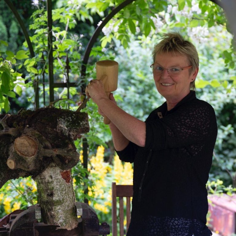 Karin Otten hält einen Holzhammer und einen Meißel, steht an einem Holzklotz im Garten.