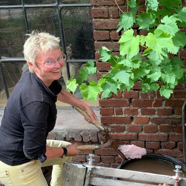 Karin Otten hält eine keramische Form in einer Zange in der Nähe einer Mauer mit Pflanzen im Hintergrund.